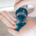 Blue gel capsules being poured from a bottle into a hand, with gloved hands handling the medication.