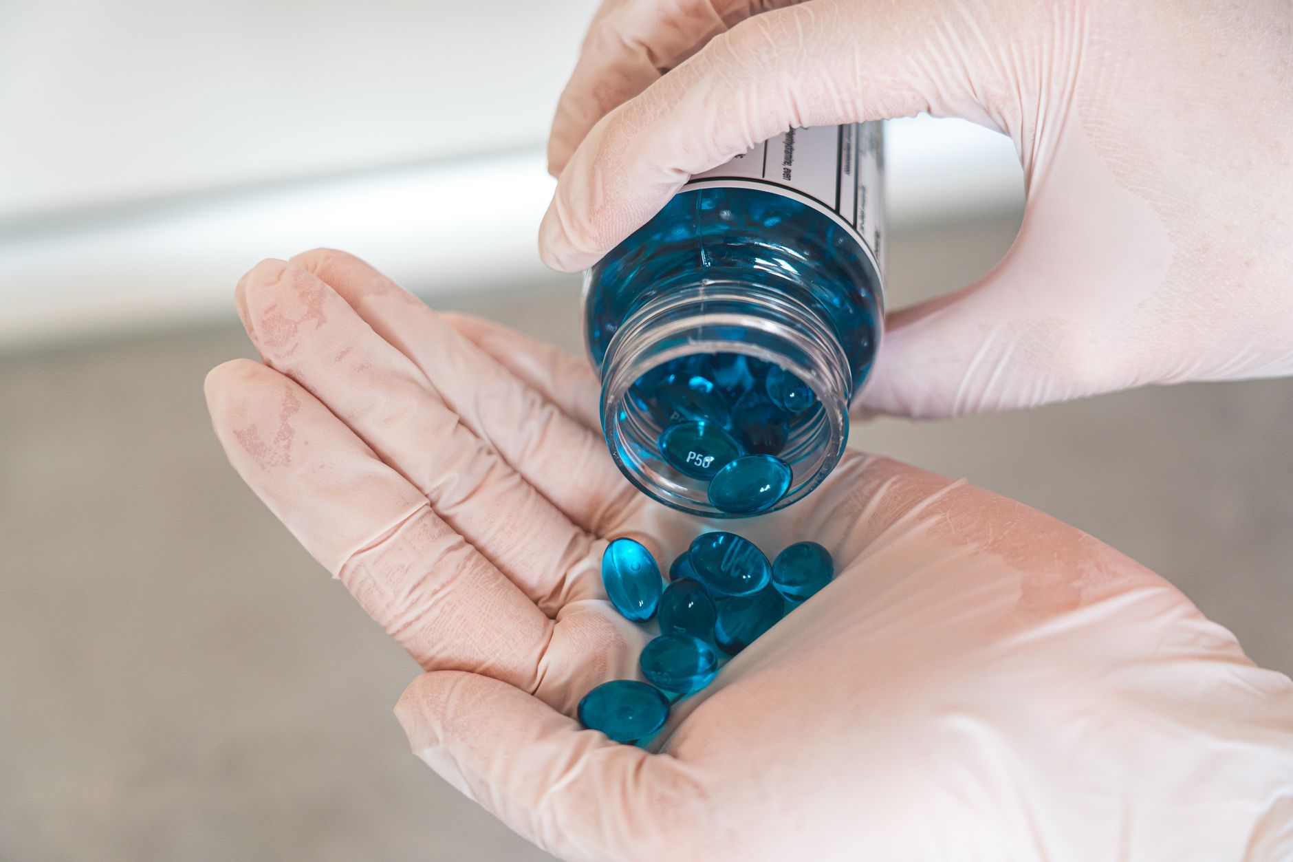 Blue gel capsules being poured from a bottle into a hand, with gloved hands handling the medication.