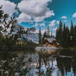 A picturesque log cabin by a tranquil lake surrounded by mountains and pine trees under a blue sky with clouds.